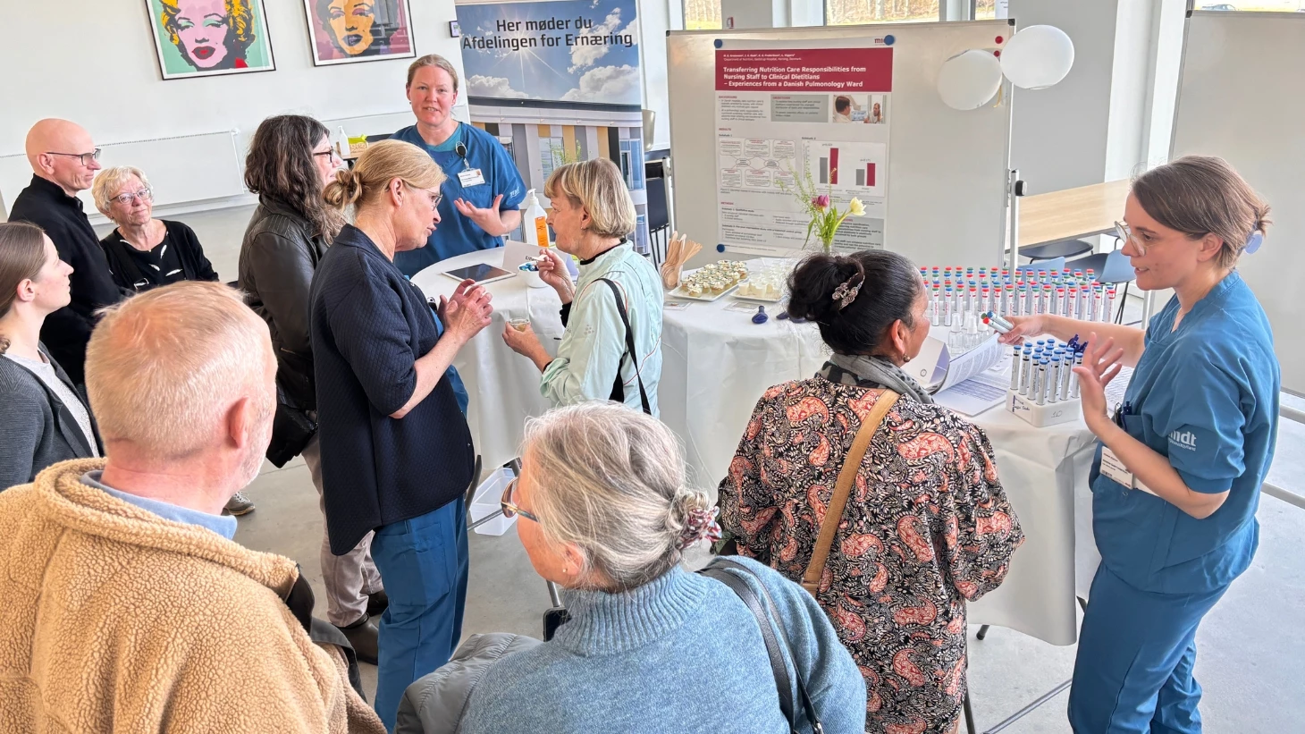 10 mennesker taler sammen om tre runde borde med hvide duge og plakater i baggrunden.