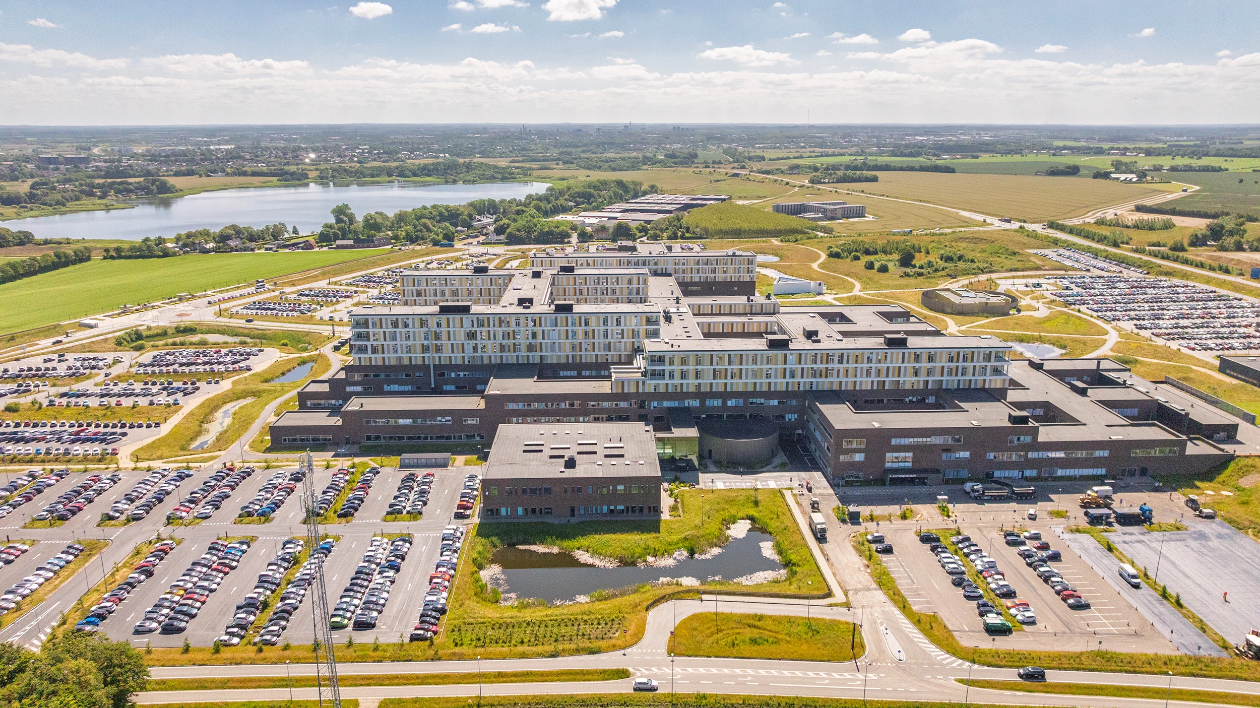 Hospital set fra oven omgivet af grønne marker og parkeringspladser. I baggrunden ses en stor sø.