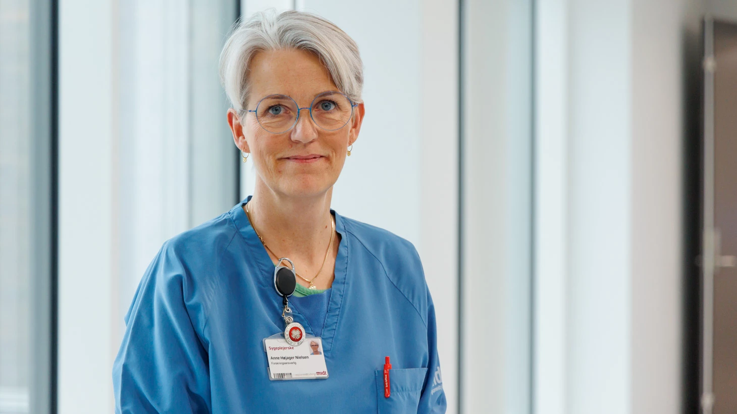 Forskningsleder Anne Højager Nielsen i blåt hospitalstøj foran store vinduer i en lys gang.