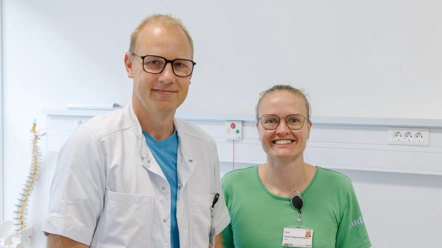 En mand og en kvinde i hospitalstøj i et klinisk miljø smiler.