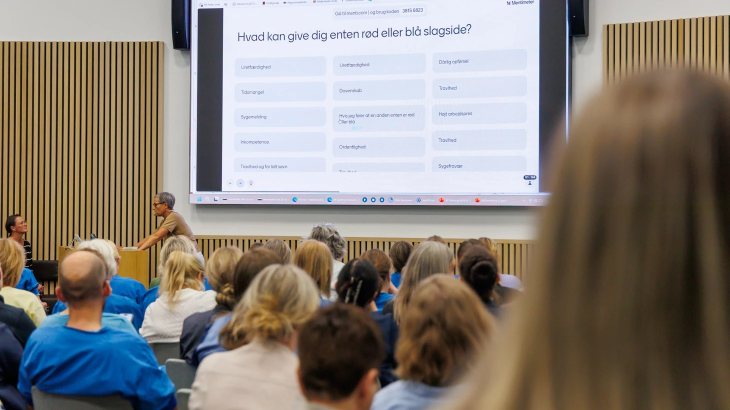Deltagere ved TRIO-workshoppen følger et oplæg om psykisk belastning i arbejdslivet. På skærmen ses spørgsmålet: ”Hvad kan give dig enten rød eller blå slagside?”