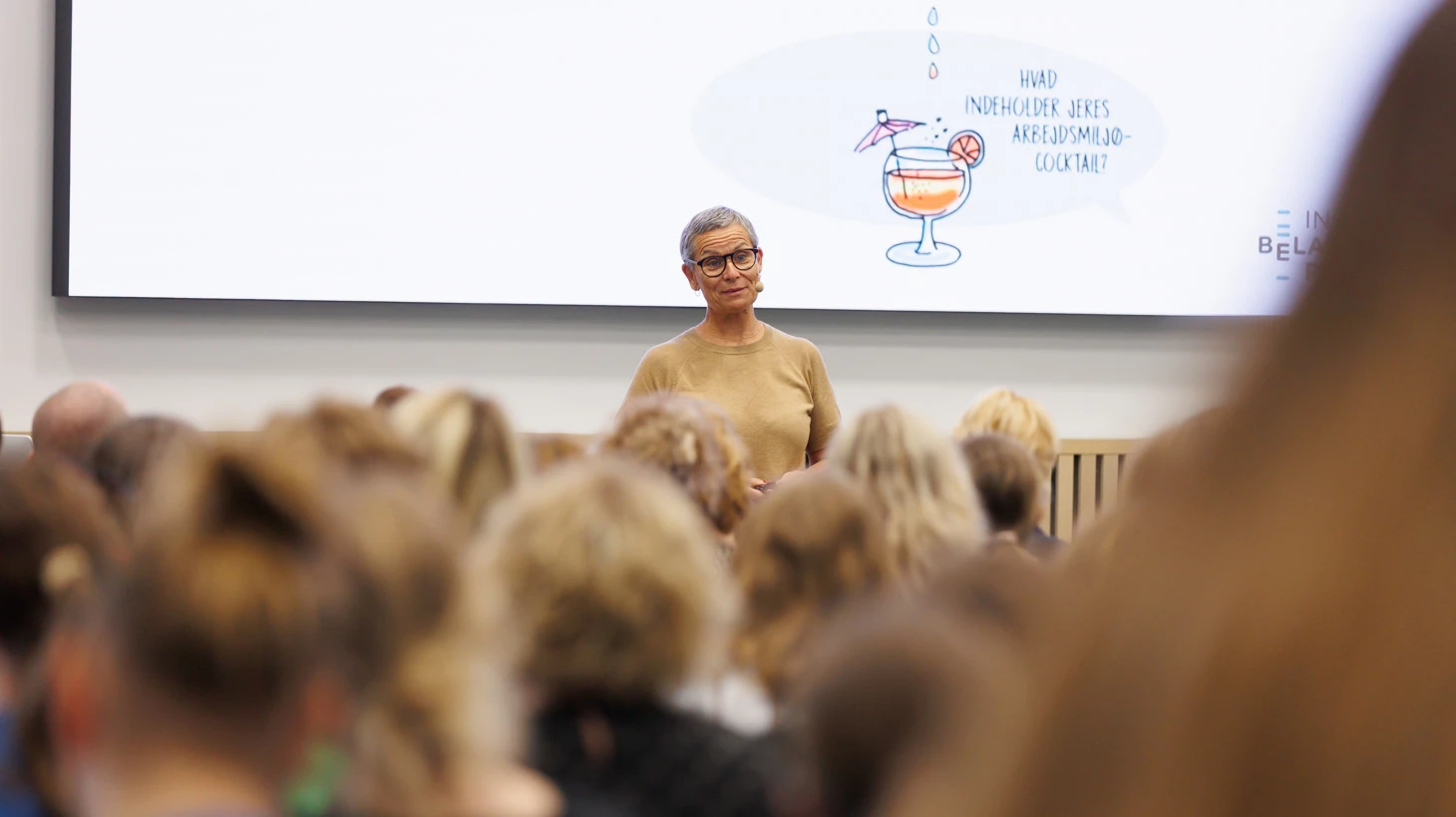 Oplægsholder står foran en stor skærm med en illustration af en cocktail og spørgsmålet ”Hvad indeholder jeres arbejdsmiljø-cocktail?” foran en sal med deltagere.