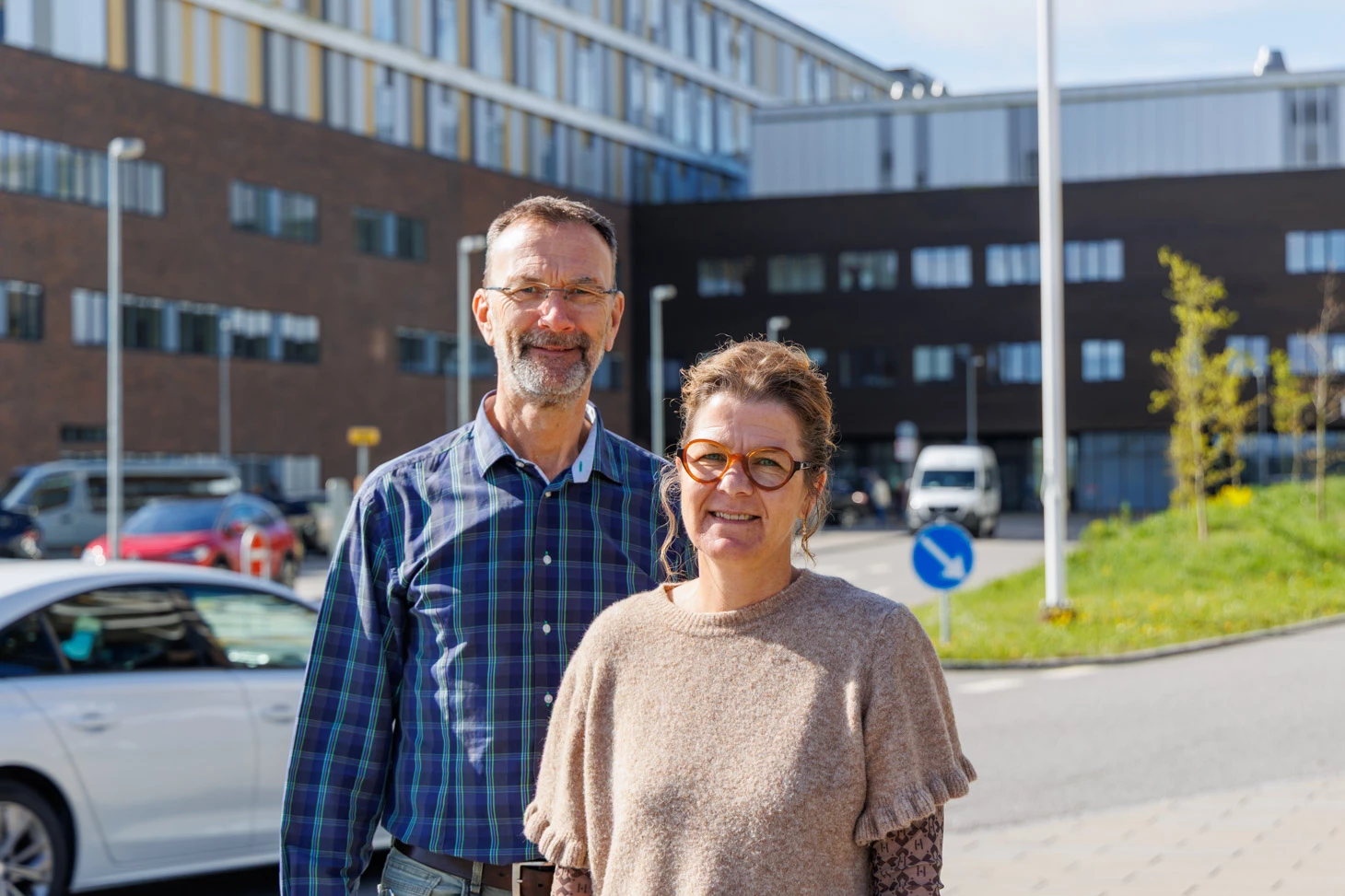 Portræt af man i blå skjorte og kvinde i lysebrun bluse foran et hospital.
