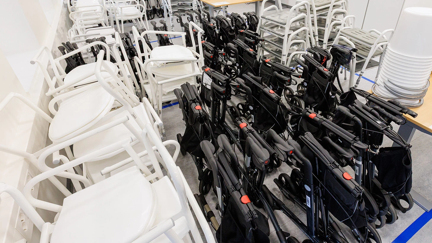 Rækker af hvide toiletforhøjere og sorte rollatorer i et depot.