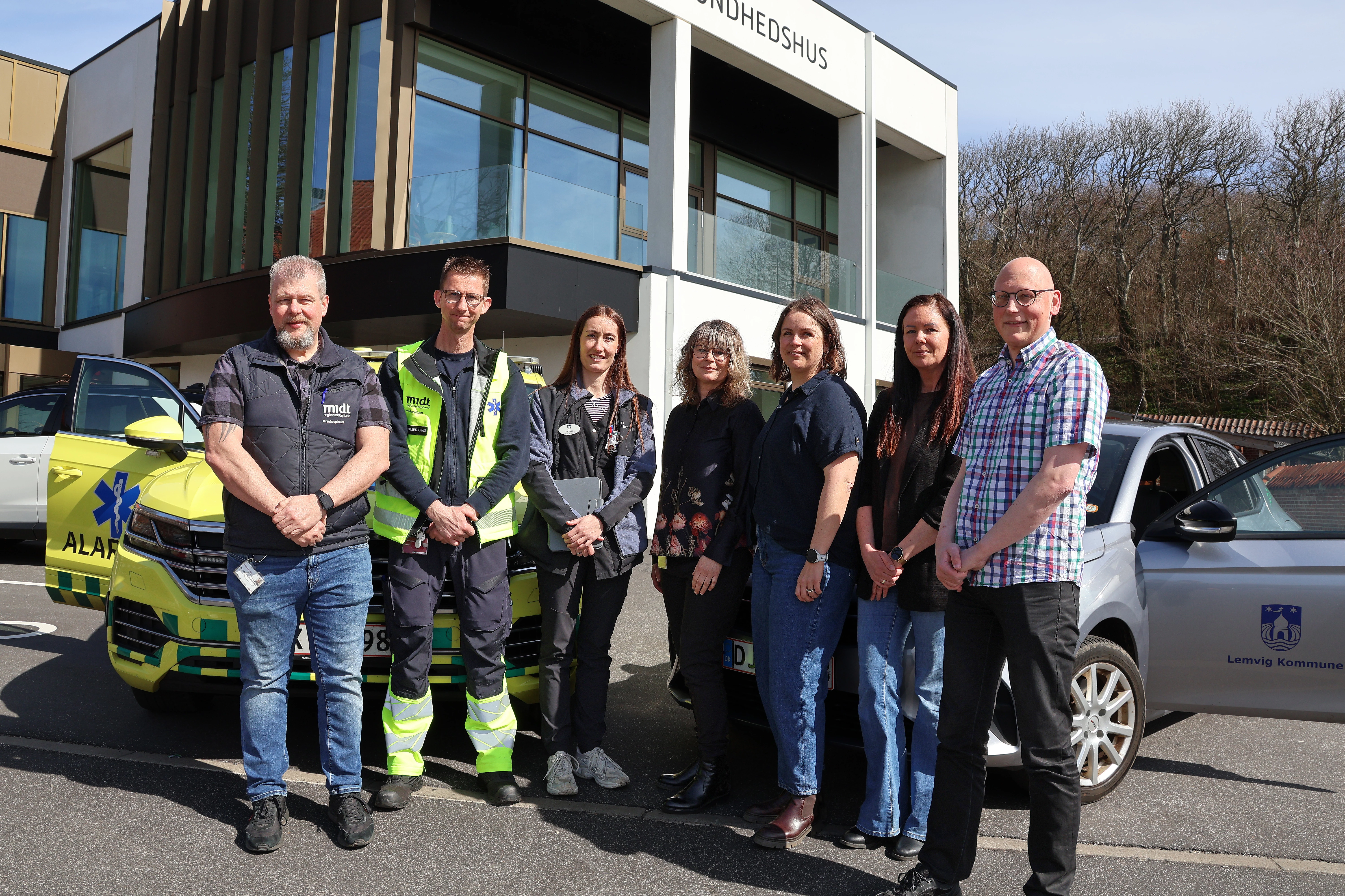 Syv personer står udenfor et sundhedshus i solskin mellem en gulgrøn akutbil og en grå bil fra Lemvig Kommune. Gruppen poser foran bygningen, og i baggrunden ses store vinduer og træer.