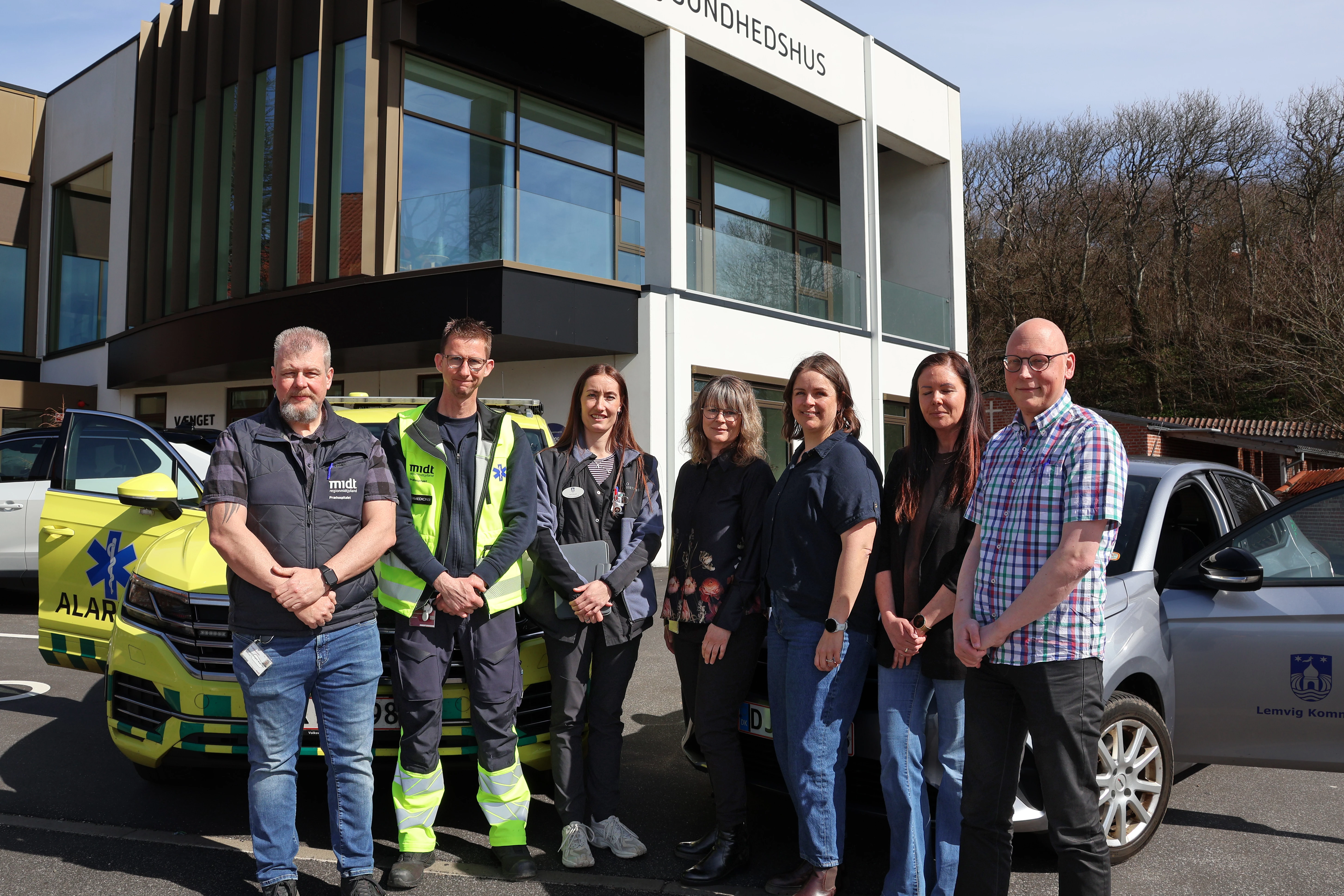 Syv personer står udenfor et sundhedshus i solskin mellem en gulgrøn akutbil og en grå bil fra Lemvig Kommune. Gruppen poser foran bygningen, og i baggrunden ses store vinduer og træer.