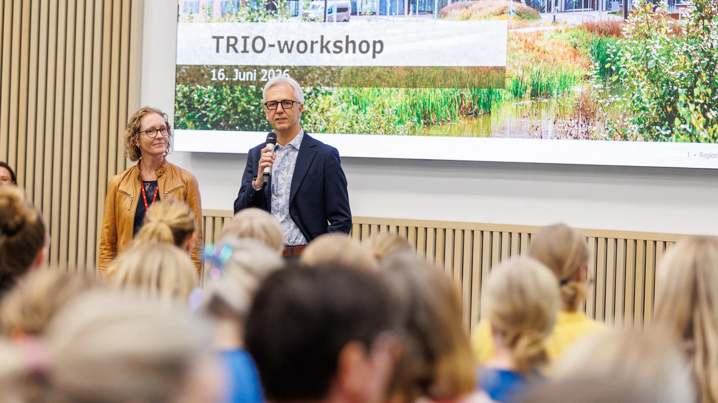 Kvinde i lyst tøj og briller og mand i mørk jakke og hvid skorte samt briller foran storskærm.