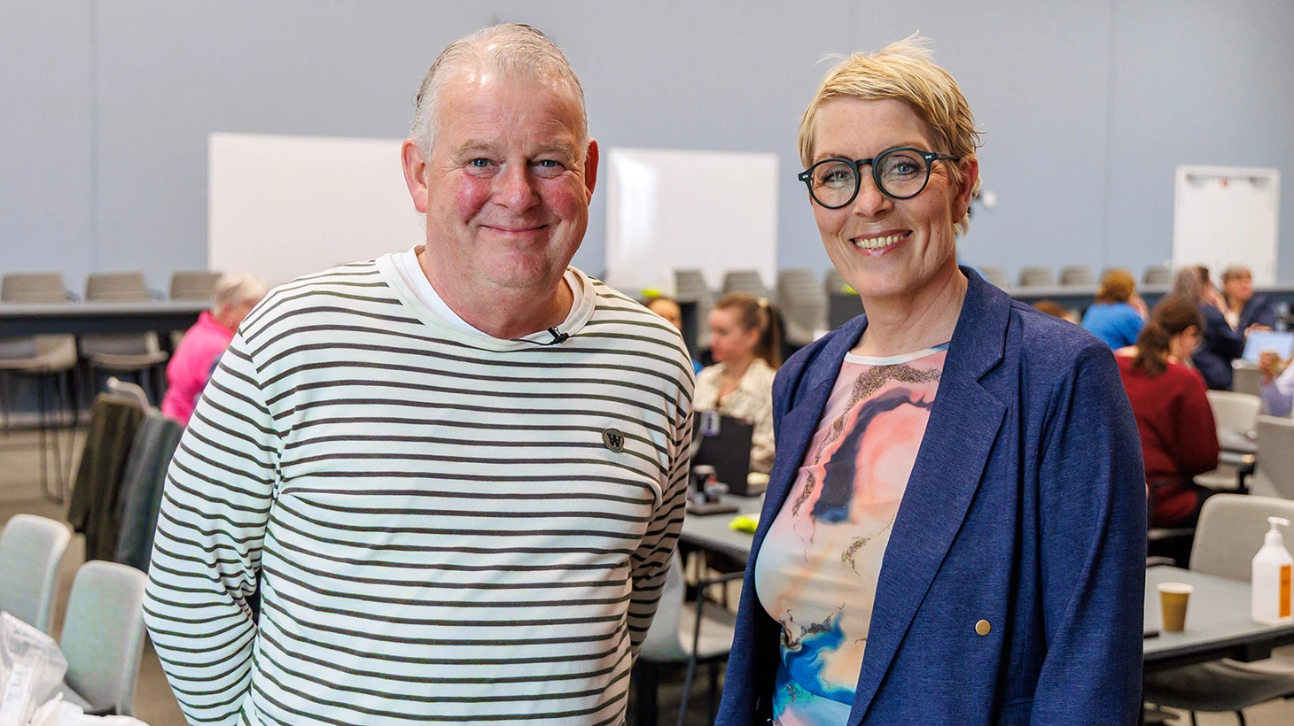 Mand i hvid og sortstribet trøje og kvinde i blå habitjakke, farvet top og briller i et undervisningslokale 